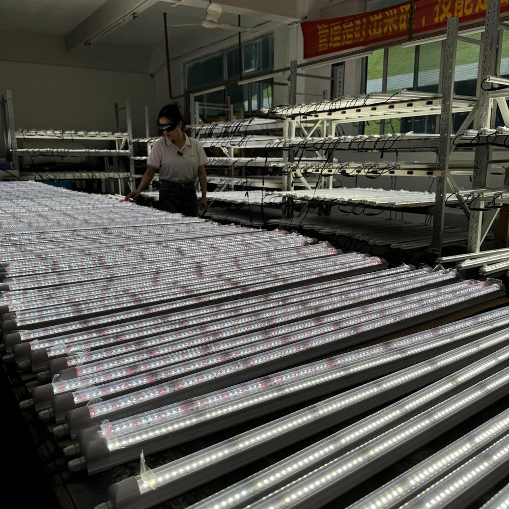 Rows of long LED grow light tube fixtures laid out on a workshop floor and on shelving during assembly and inspection, with a worker checking a batch in the background